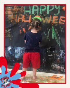 Child with hair in bunches, wearing a navy top and red shorts, standing barefoot in front of a painted ‘Happy Halloween’ mural at a messy play session
