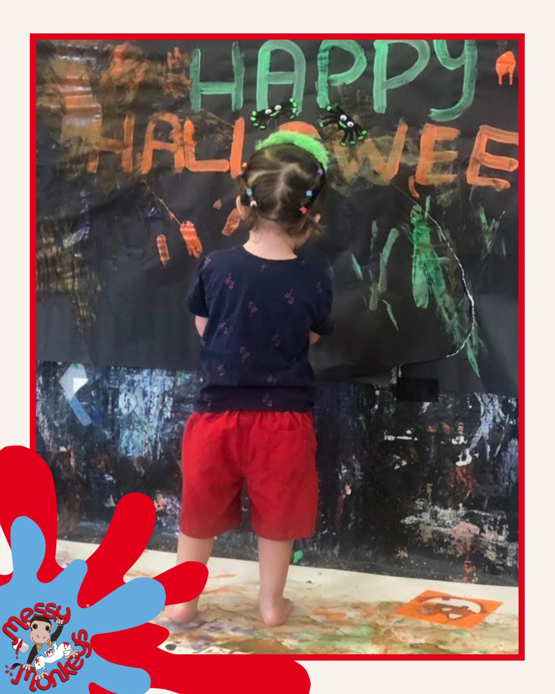 Child with hair in bunches, wearing a navy top and red shorts, standing barefoot in front of a painted ‘Happy Halloween’ mural at a messy play session
